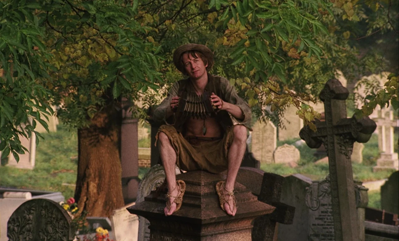 Mahler (1974) movie still: fullbody — A rural cemetery features numerous aged stone headstones scattered beneath a canopy of trees. A thin man in…