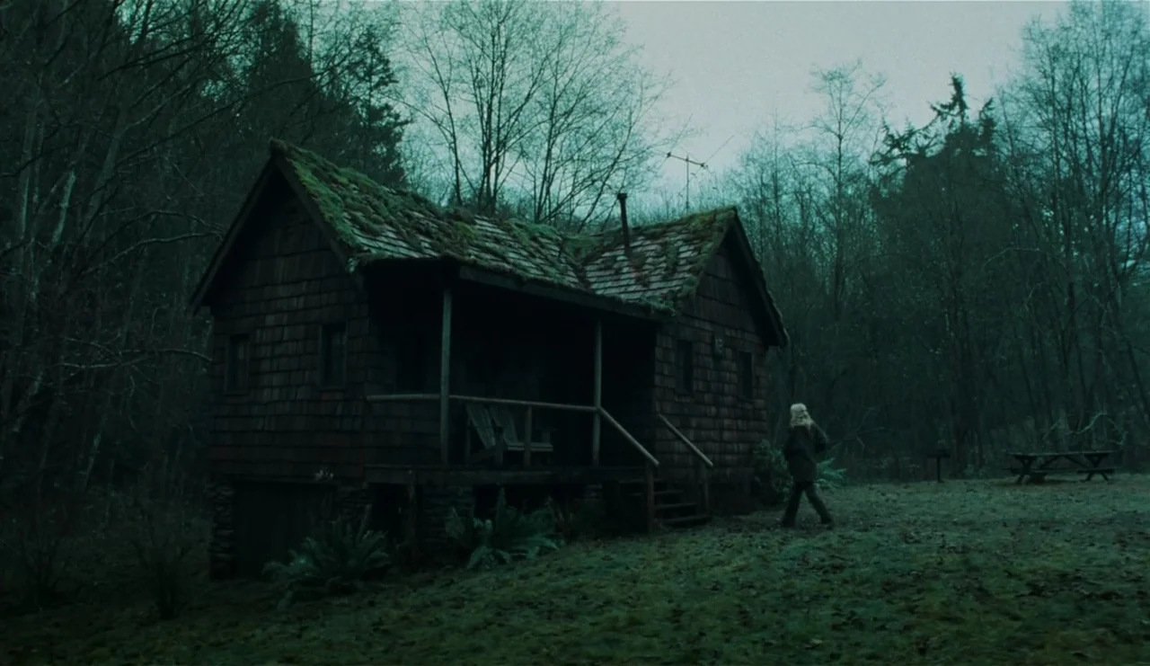 The Ring (2002) movie still: wide — A weathered wooden cabin with a moss-covered roof sits in a dense evergreen forest. A woman in dark clothing…
