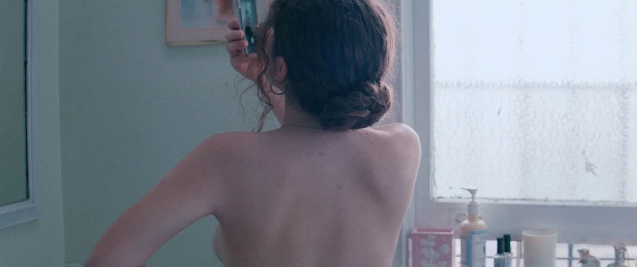 Shiva Baby (2020) movie still: medium — A light-toned bathroom features a frosted glass window and a vanity surface. A young woman turns away from…
