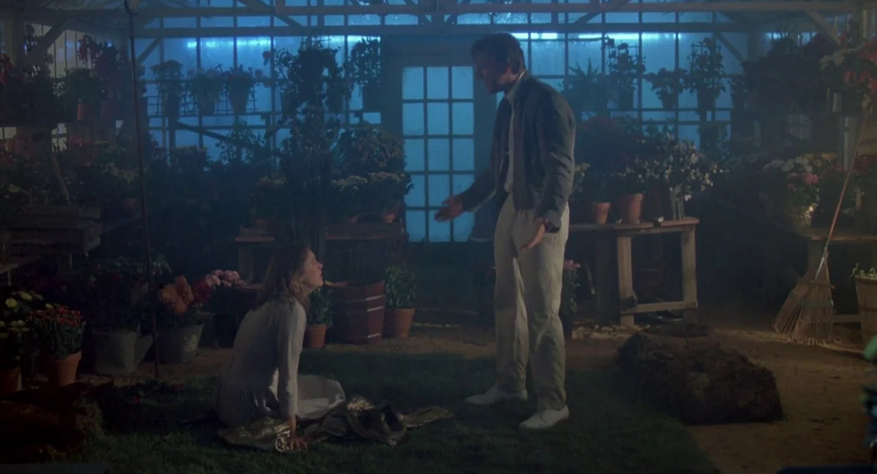 Peggy Sue Got Married (1986) movie still: fullbody — A glass-walled greenhouse contains rows of potted flowers and greenery under dim blue light. A…