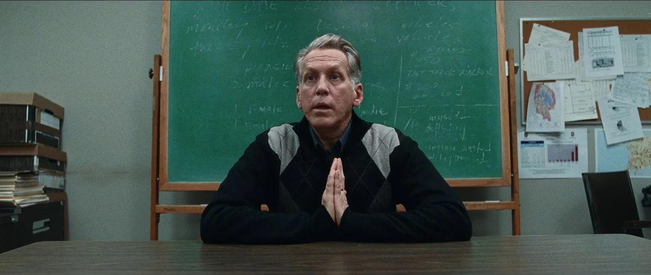 Bad Education (2019) movie still: medium — A man sits at a wooden table in front of a green chalkboard. The chalkboard is filled with handwritten…