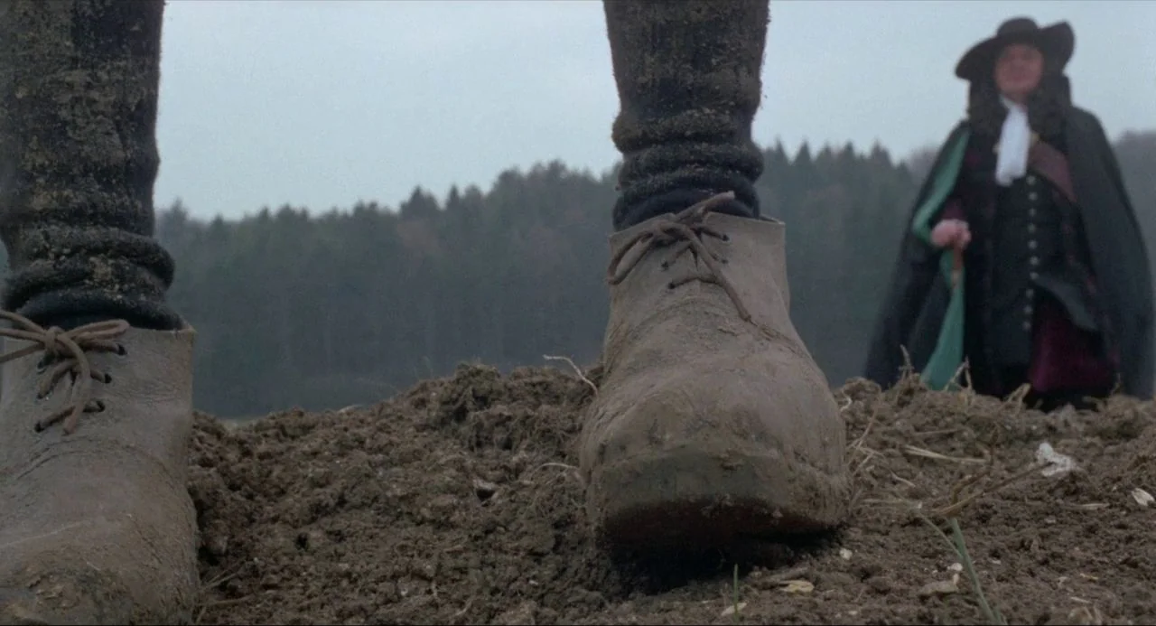 The Blood on Satan’s Claw (1971) movie still: closeup — A field of ploughed earth features muddy boots in the immediate foreground. In the distant…