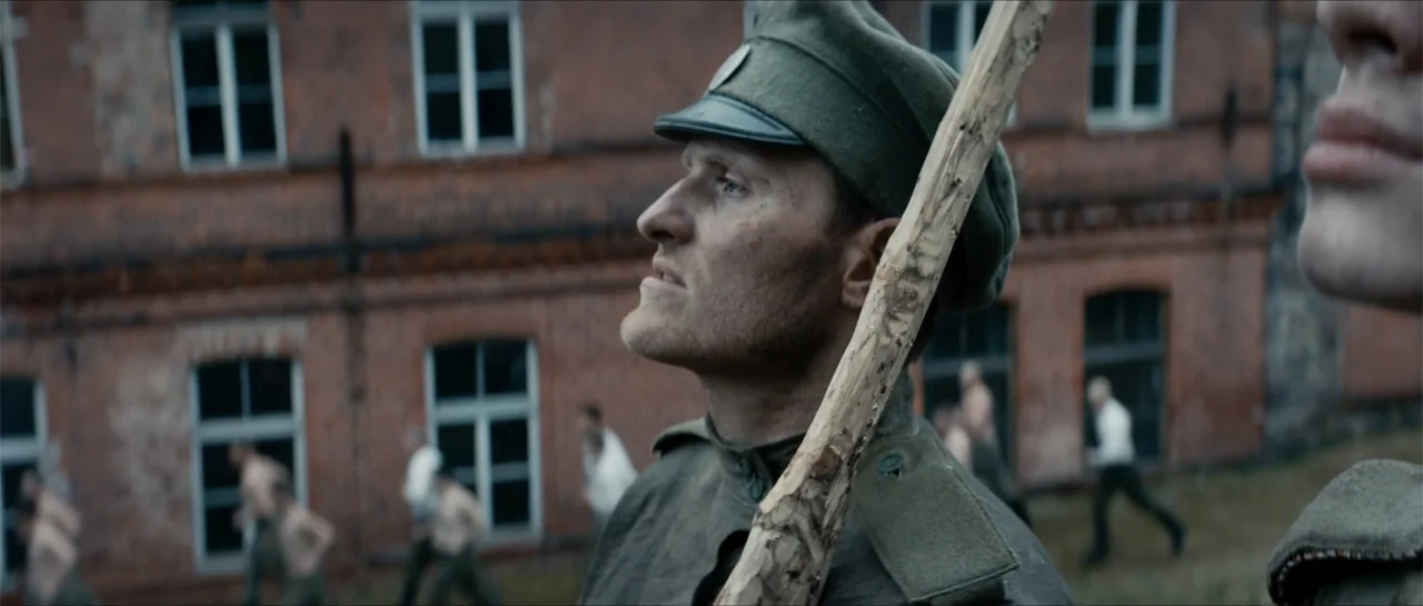 The Rifleman (2019) movie still: medium — A red brick military building stands behind a field with soldiers running around it. A soldier in the…