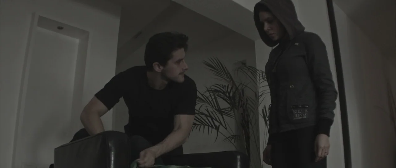 Amor Mutante (2019) movie still: medium — A modern interior space features white walls and a large potted palm plant. A young man sitting in a dark…