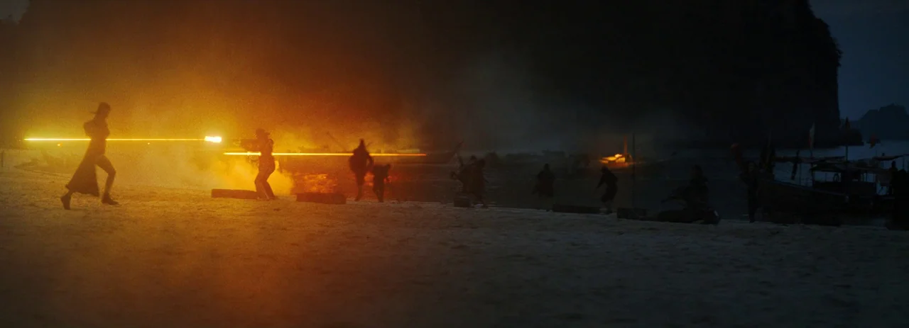The Creator (2023) movie still: wide — A dark night beach scene features silhouettes of people moving across sandy terrain. A bright glowing laser…