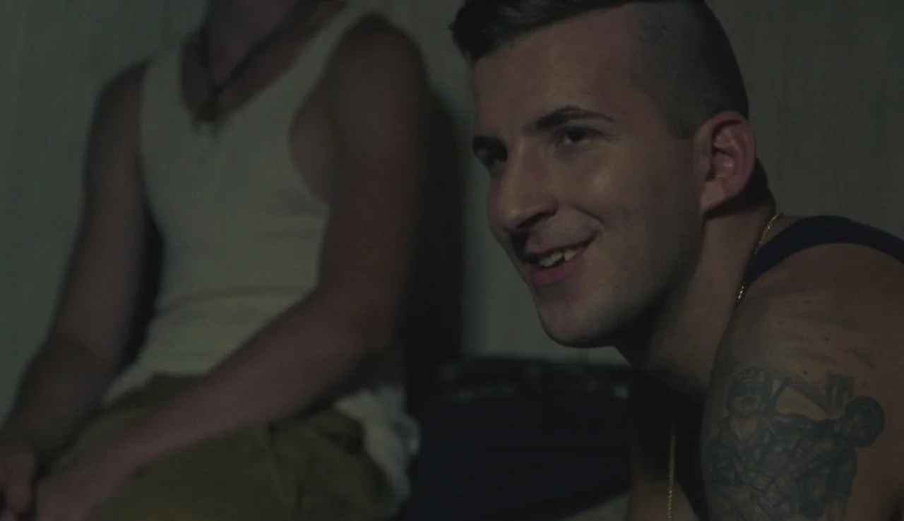 Beach Rats (2017) movie still: medium — A dimly lit interior space contains two young men sitting close to each other. The primary subject in the…
