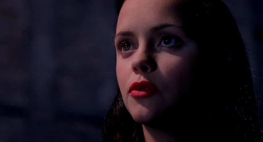 The Man Who Cried (2000) movie still: closeup — A dark, dimly lit indoor space with textured walls hidden in deep shadow. A young woman with dark…