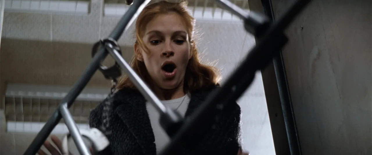 Conspiracy Theory (1997) movie still: medium — An interior staircase with metal railing and fluorescent overhead lighting fixtures. A woman looks…