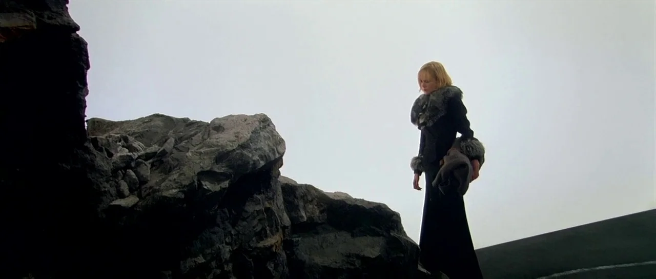 Dogville (2003) movie still: fullbody — A woman in a black coat stands on a dark, jagged rock formation. An expansive, featureless light-grey sky…
