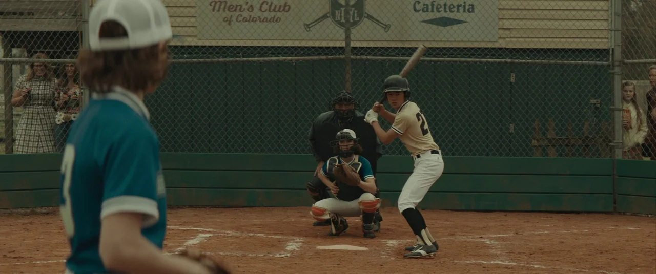 The Black Phone (2021) movie still: medium — A baseball game plays on a dirt field in front of a chain-link fence. A young batter in a tan uniform…