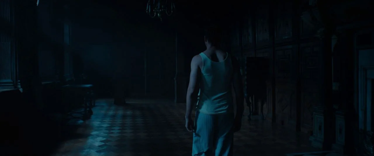Rebecca movie still: fullbody — A dark, wood-paneled corridor features a checkered marble floor and an overhead chandelier. A man in sleepwear…