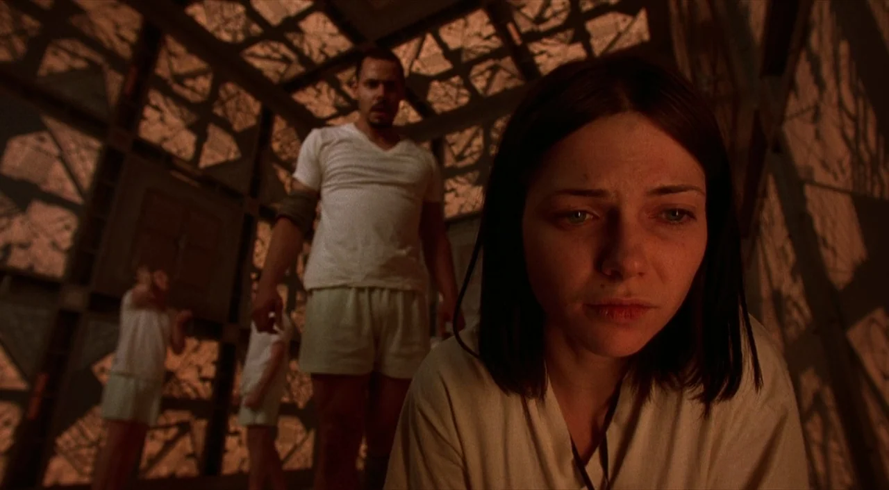 Cube (1997) movie still: closeup — A square cell is constructed of interlocking metal grates and panels with patterned shadows. A young woman stands…
