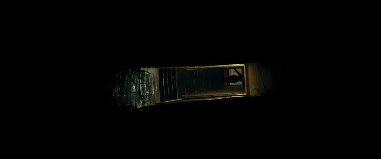 Saint Maud (2019) movie still: establishing — A dark stairwell viewed through a small, narrow horizontal slit in the wall. The stairwell leads down…
