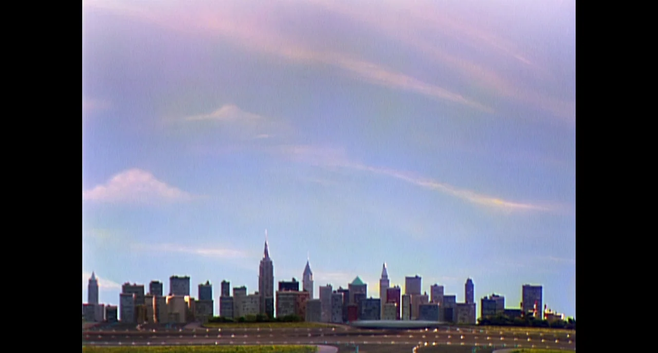 A Beautiful Day In The Neighborhood movie still: establishing — A wide view shows a miniature-style city skyline set against a pale blue sky. The…