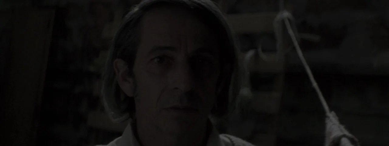 Post Mortem (2010) movie still: closeup — A dark, dimly lit indoor space with indistinct textures visible in the deep shadows. A middle-aged man…
