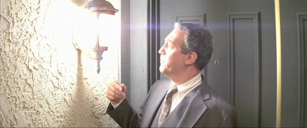 Poltergeist movie still: medium — A man stands near a front door with textured stucco walls. He looks at a bright exterior light fixture mounted on…