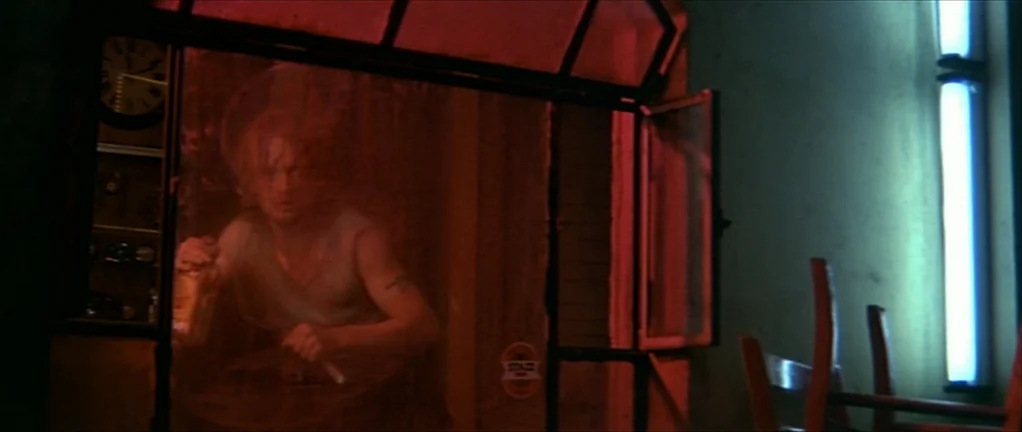 The Moon in the Gutter (1983) movie still: medium — A dimly lit bar area features a person standing behind a glass partition wall. A man in a…