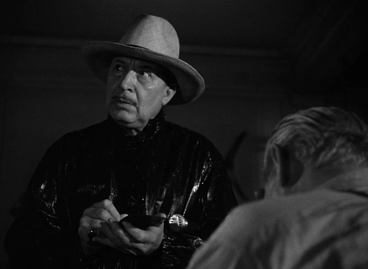Key Largo (1948) movie still: medium — A dimly lit interior space with deep shadows and high-contrast lighting. A man wearing a hat stands in the…