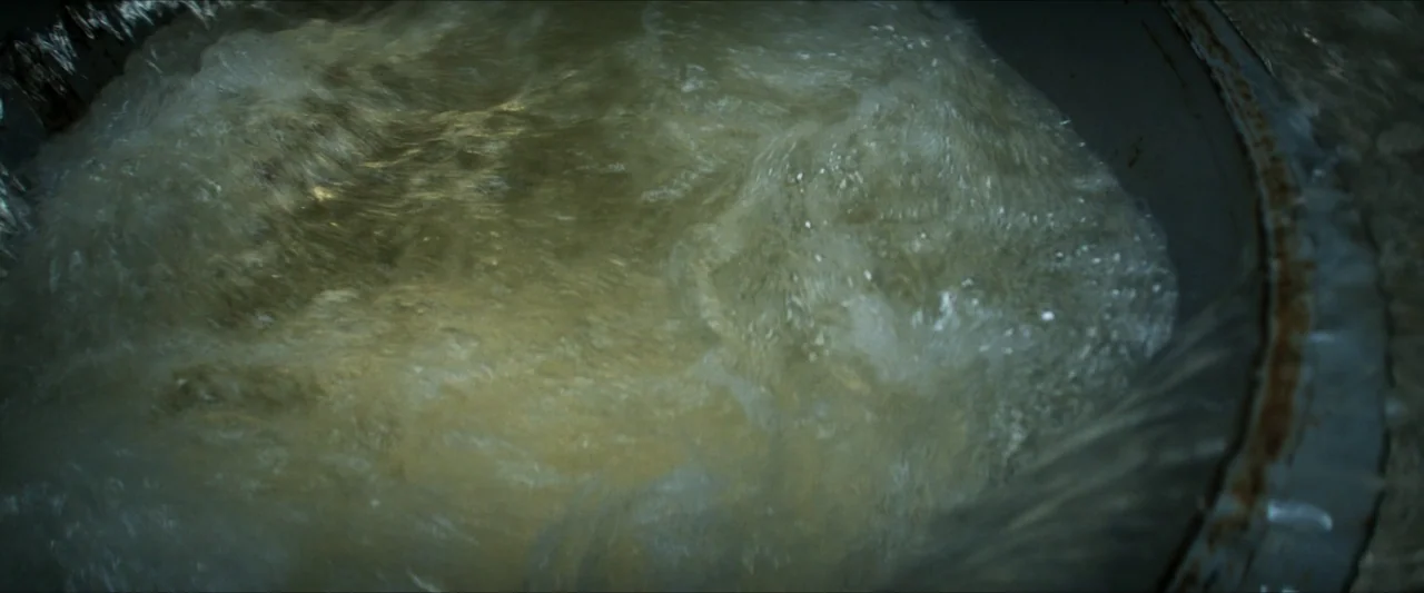 Last Breath (2025) movie still: closeup — A close view of opaque liquid swirling in a circular metal basin drain. The turbulent water rotates…