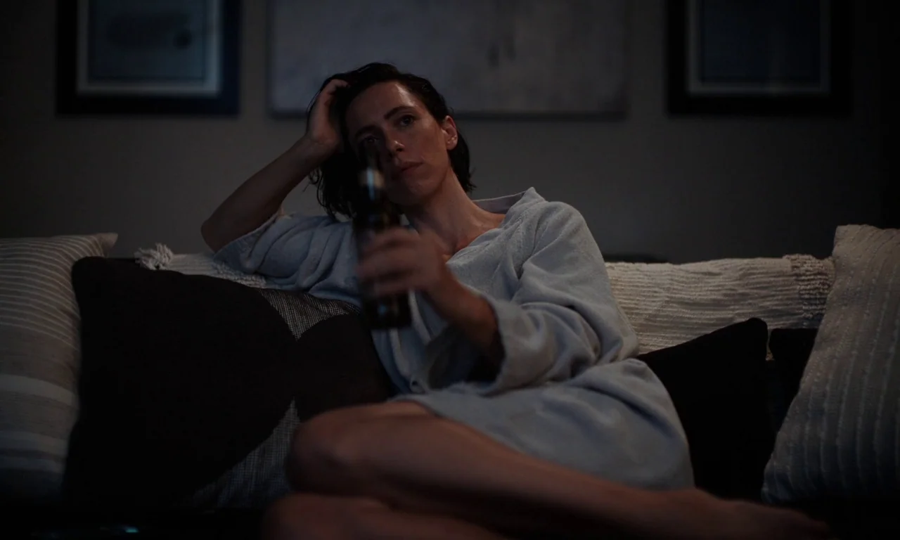 Resurrection (2022) movie still: medium — A dark, dimly lit room contains a person resting on a couch. The person holds a bottle while looking away…