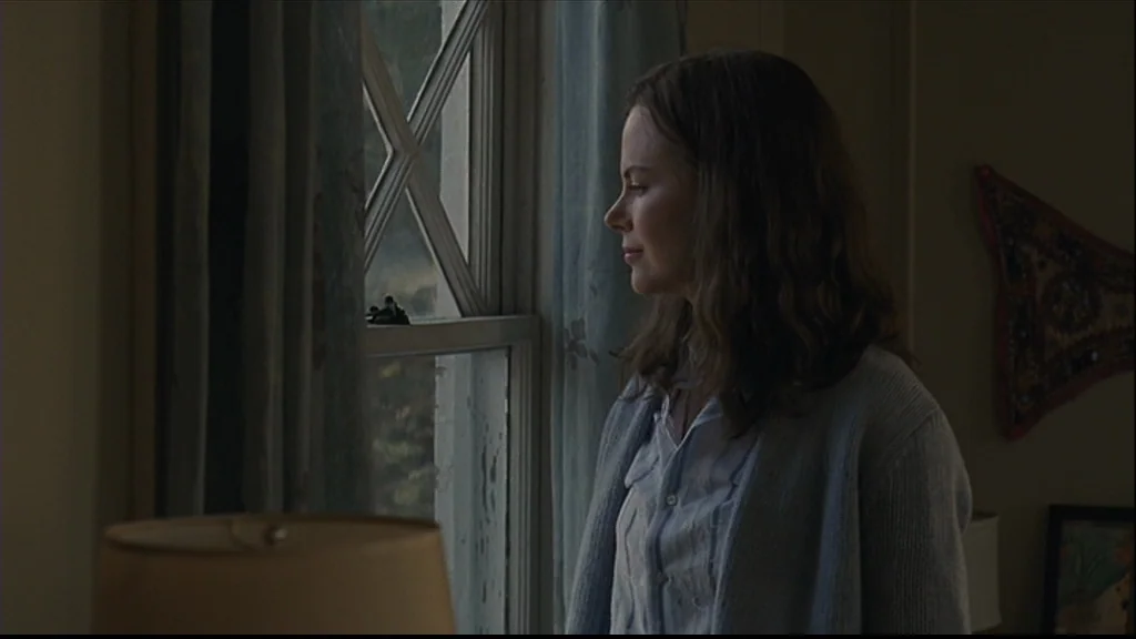 Margot At The Wedding (2007) movie still: medium — A domestic interior with beige walls, sheer blue curtains, and a window frame. A woman with light…