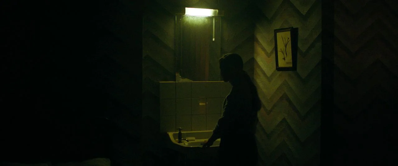 Saint Maud (2019) movie still: medium — A dimly lit bathroom features chevron-patterned wallpaper and a tiled sink area. A person stands facing the…