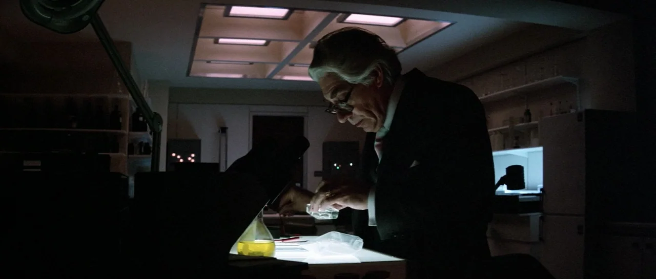 Lifeforce (1985) movie still: medium — A dimly lit scientific workspace with metal shelves and recessed ceiling panels. An older man in a suit…