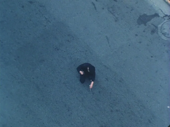 Trial (1975) movie still: wide — A man in dark clothing kneels on a vast expanse of grey asphalt. He touches an object to the textured road surface…