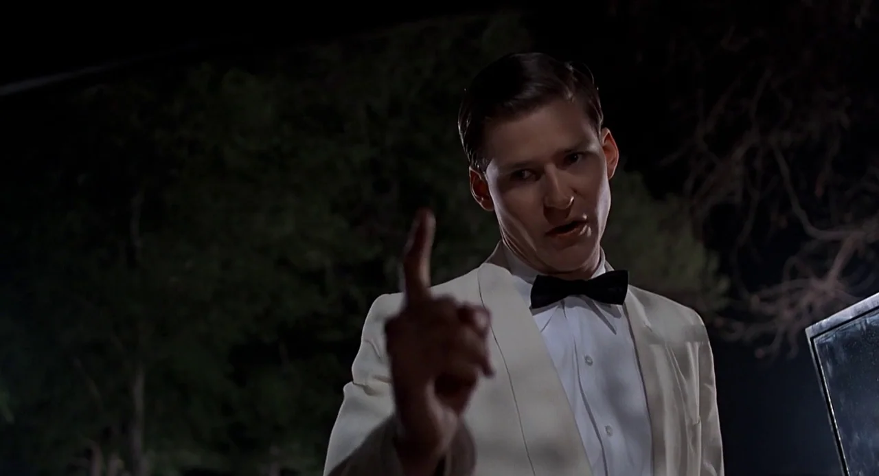 Back to the Future (1985) movie still: medium — A man in a tuxedo stands outdoors at night in front of dark trees. The man points his index finger…