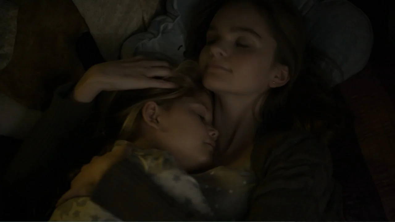 Totem (2022) movie still: closeup — A dimly lit bed with dark bedding and soft textures. A young woman holds a sleeping child closely in her arms.