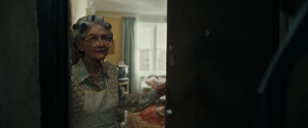 We Are Zombies (2023) movie still: medium — An elderly woman stands in a doorway of a home with a patterned rug. She is opening the dark wooden door…