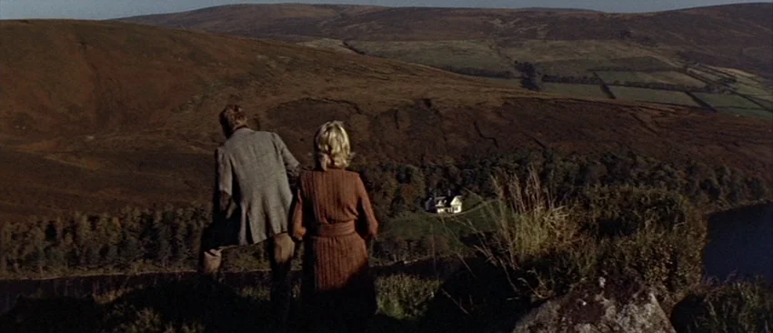 Images (1972) movie still: wide — A man and woman overlook a vast, hilly landscape of brown and green terrain. A small isolated house sits in the…