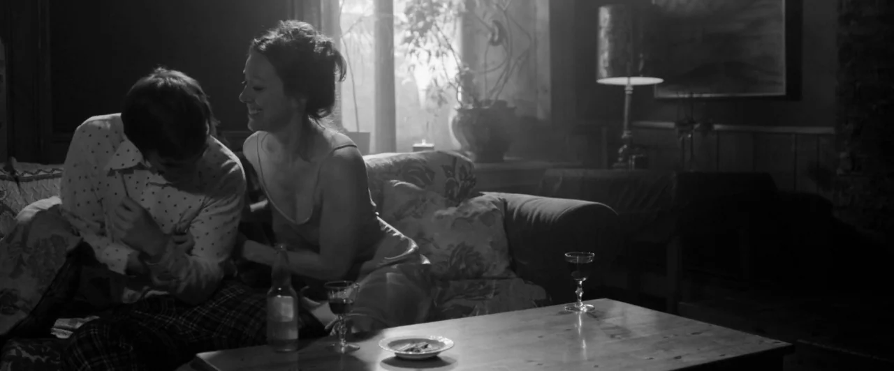 Weirdos (2016) movie still: medium — A dimly lit room contains a patterned sofa and a low wooden coffee table. A man and a woman sit closely on the…
