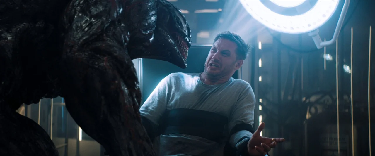 Venom (2018) movie still: medium — A clinical, modern research facility features metallic surfaces and bright medical lights above. A man restrained…