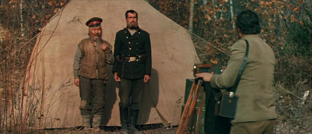 Dersu Uzala (1975) movie still: fullbody — A woodland clearing features a large canvas tent pitched amongst autumn trees and leaves. Two men stand…