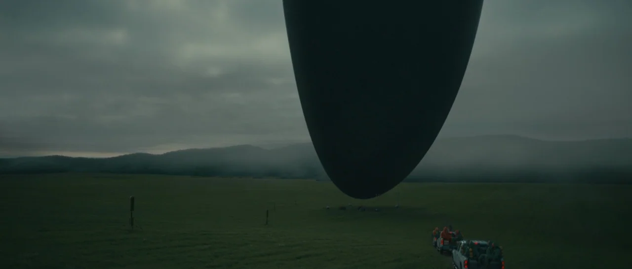 Arrival (2016) movie still: wide — A sprawling green pasture sits beneath a cloudy sky and rolling distant hills. A colossal, dark, teardrop-shaped…