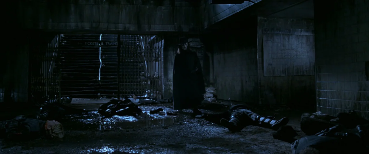 V for Vendetta (2006) movie still: wide — A dimly lit, grimy subway station platform features tiled walls and metal gates. A cloaked figure stands…