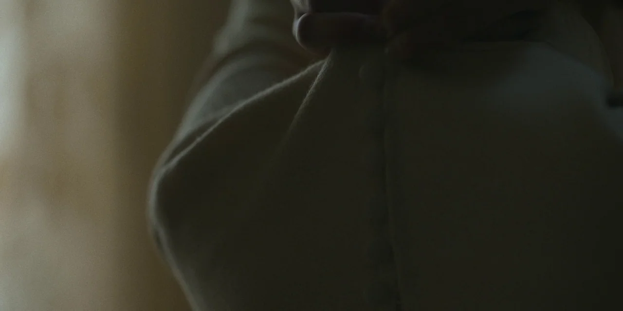 Immaculate (2024) movie still: closeup — A close view of fabric texture showing hands holding a light-colored buttoned garment. The surface of the…