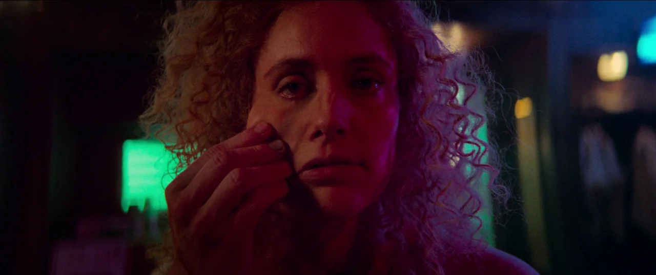 White Noise (2022) movie still: closeup — A dimly lit interior space features high contrast magenta and green neon lighting. A woman with curly…