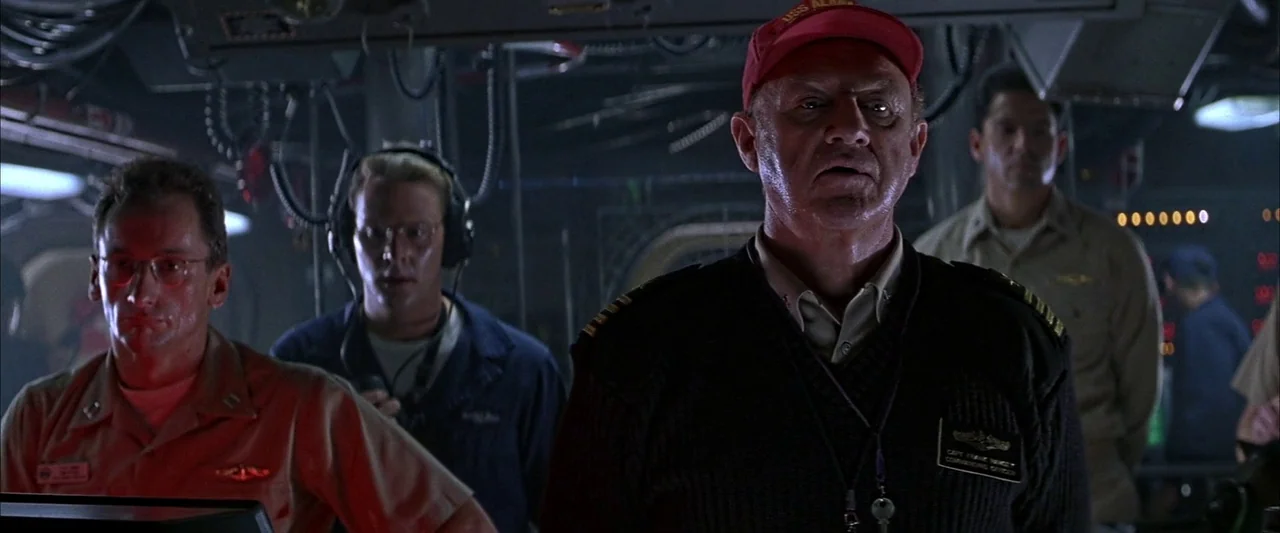 Crimson Tide (1995) movie still: medium — A dark, cramped control room filled with wires, consoles, and dimly lit metallic surfaces. A commanding…