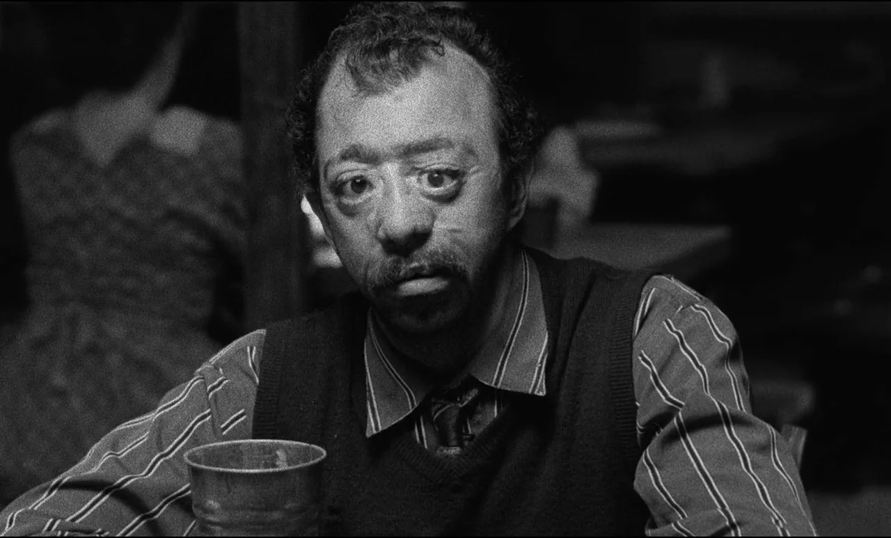 Go Down Death movie still: medium — A dimly lit indoor space with a blurred person visible in the background. A man sits at a table and looks…