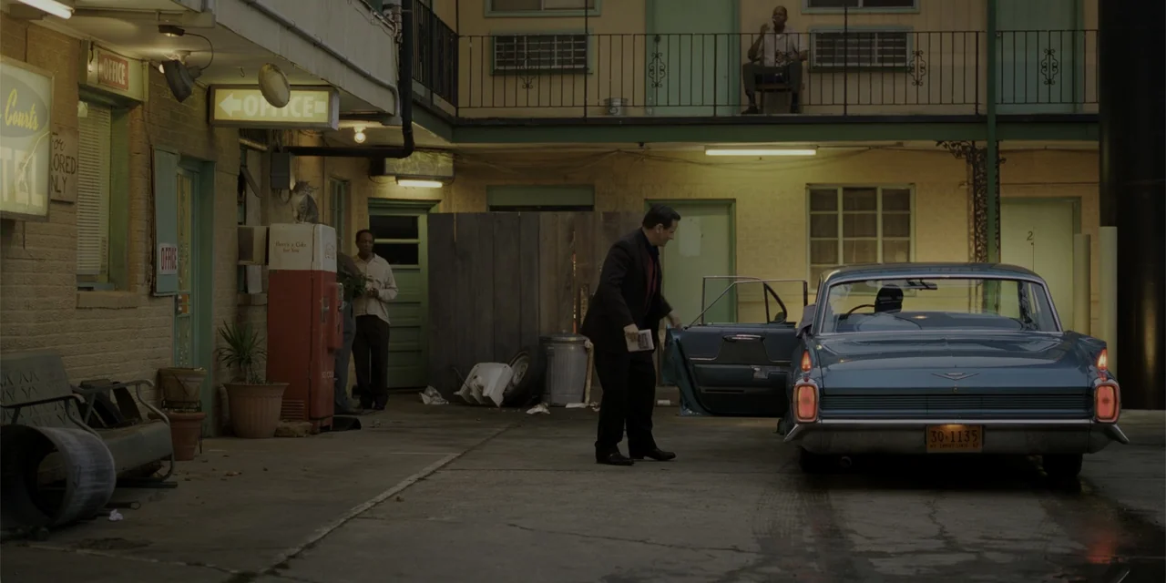Green Book (2018) movie still: wide — A 1960s-style motel exterior features a blue vintage Cadillac parked in the courtyard. A man exits the vehicle…