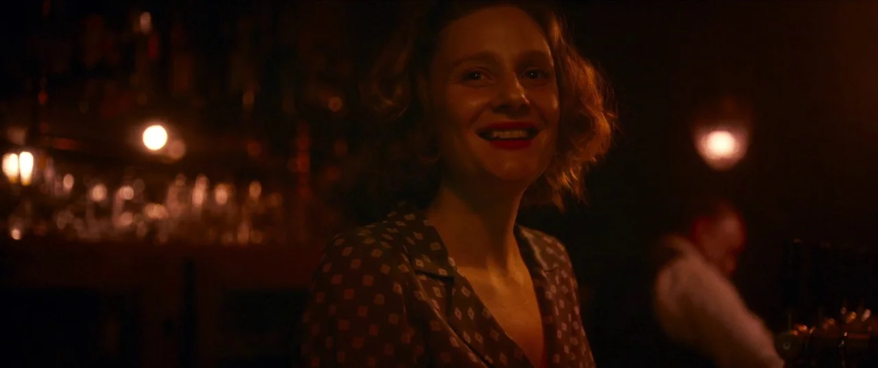 Earwig (2022) movie still: medium — A dimly lit room features a bar area with blurry illuminated glass objects behind. The woman wears a dark…