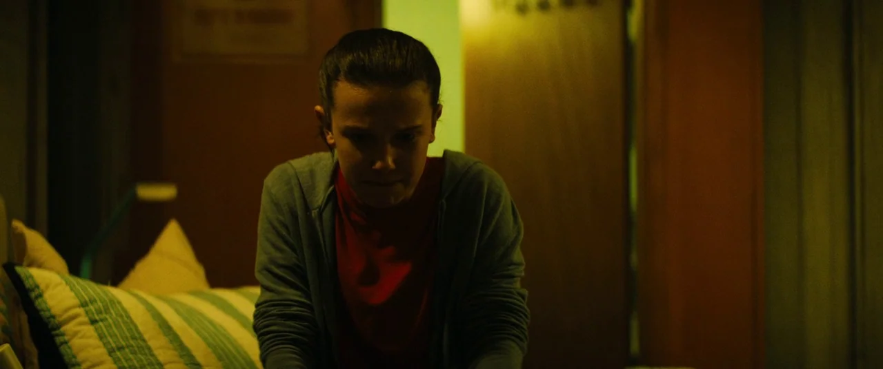 Godzilla: King of the Monsters (2019) movie still: medium — A dimly lit room with yellow walls and visible pillows in the foreground. A young woman…