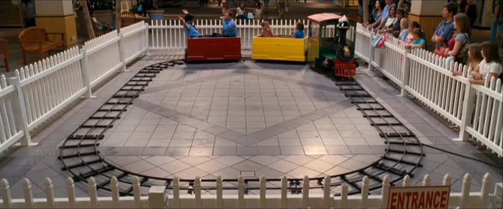 Observe And Report (2009) movie still: wide — A miniature train track sits inside a fenced enclosure within a large shopping mall. Children and…