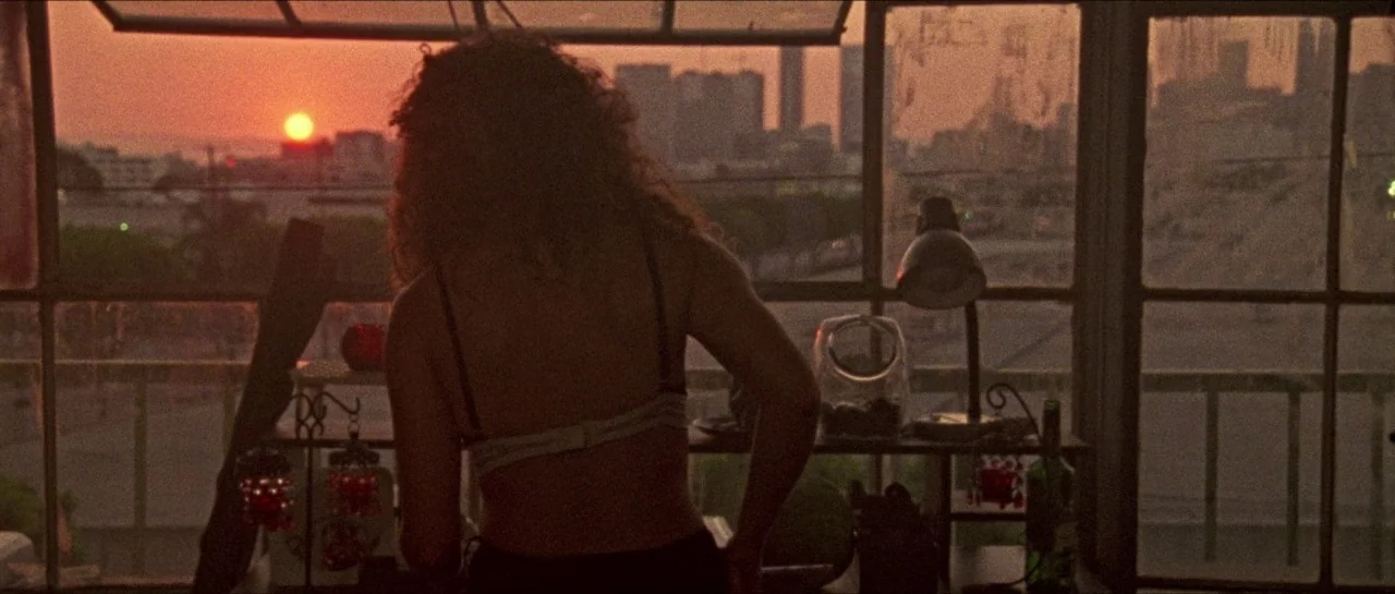 Bliss movie still: medium — A woman stands in a dim apartment interior facing a large multi-paned window. Outside the window a city skyline is…