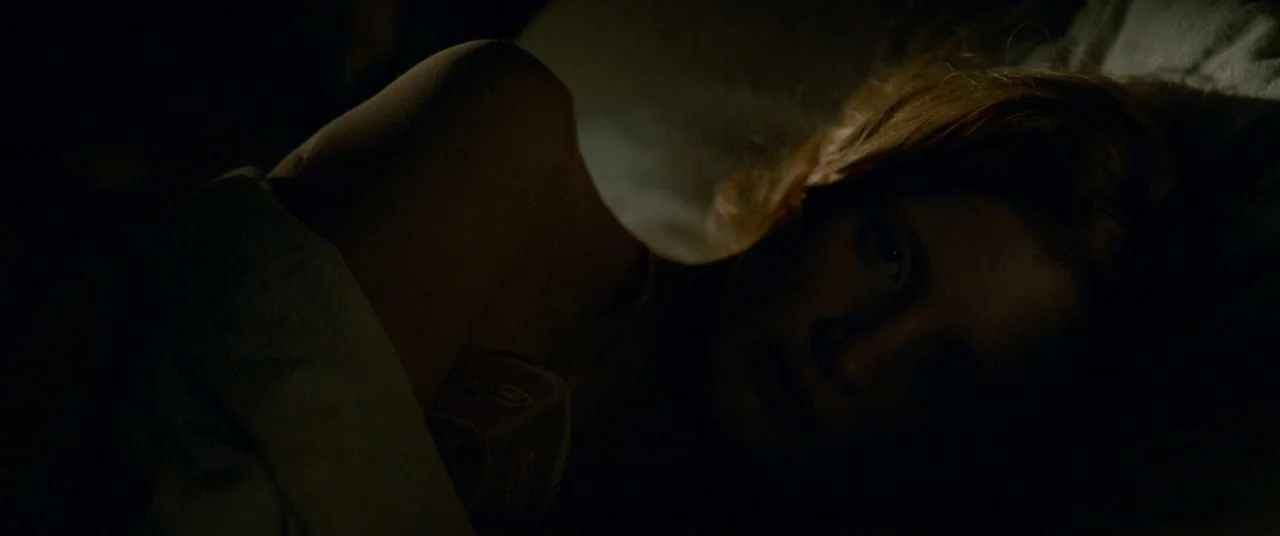 Lawless (2012) movie still: closeup — A dimly lit room at night with a person resting in bed. The subject lies under white covers with her face…