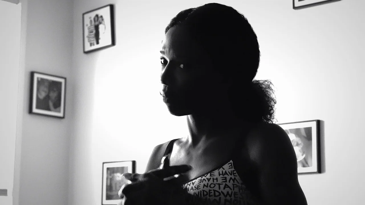 Kokomo City (2023) movie still: medium — A light-walled room interior features several framed rectangular artworks hanging on the wall. A woman with…