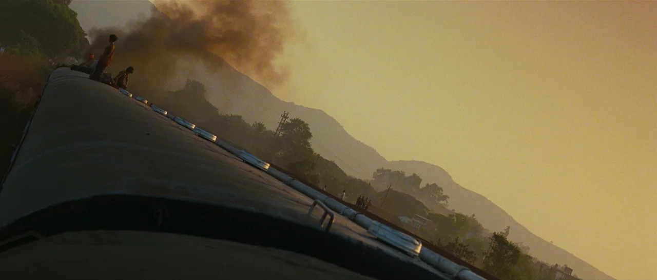 Slumdog Millionaire (2008) movie still: wide — A group of people sits on top of a moving train during sunset. Mountains rise in the background under…