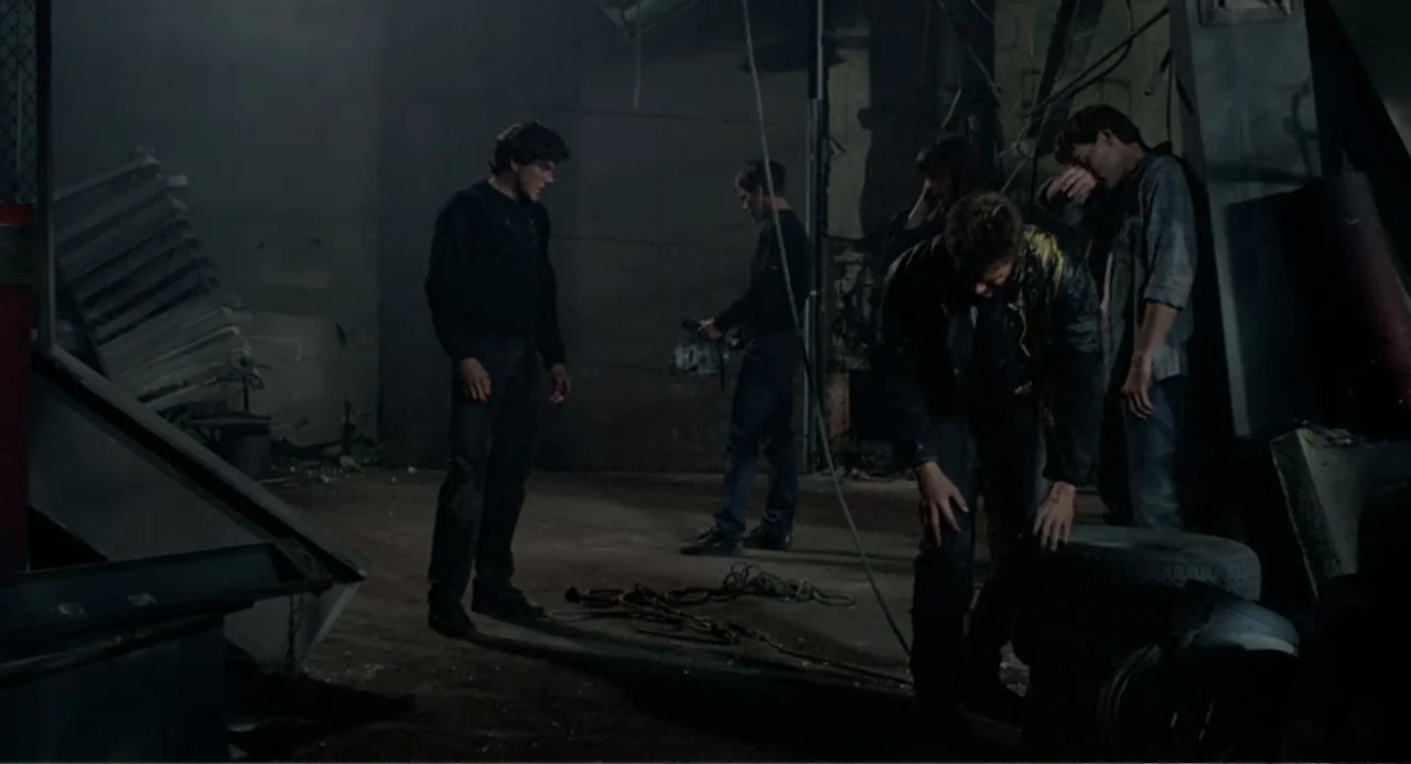 Dangerously Close (1986) movie still: wide — A dimly lit industrial space contains scattered rubble and discarded car tires. Four young men stand in…